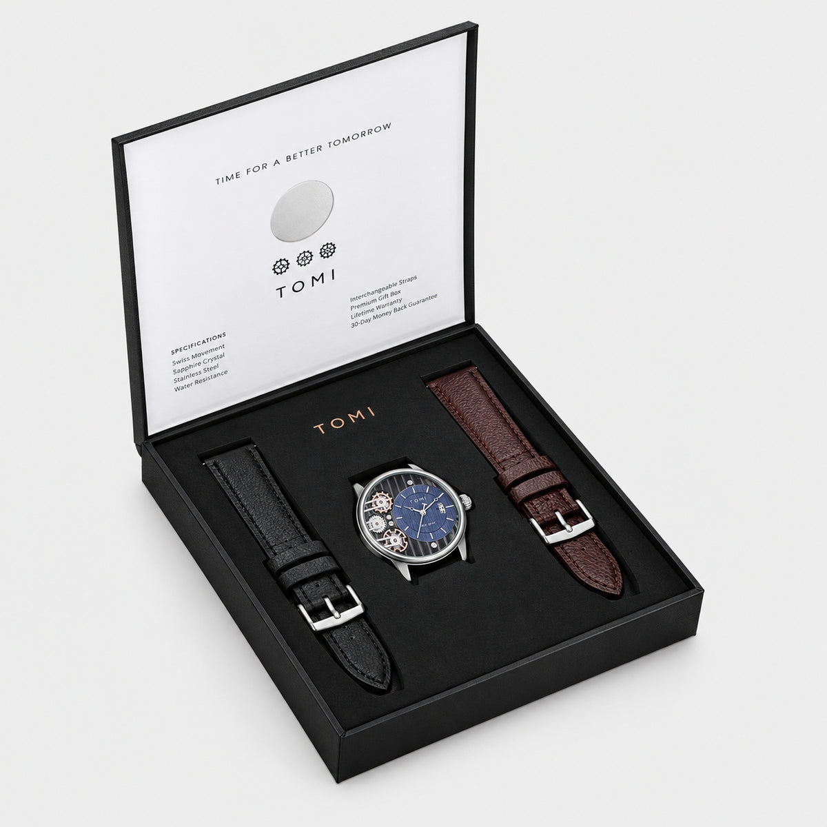 A TOMI watch gift set in an open black presentation box on a light gray background. The set includes a silver-toned watch with a blue dial and skeletonized movement, a black leather strap, and a brown leather strap.
