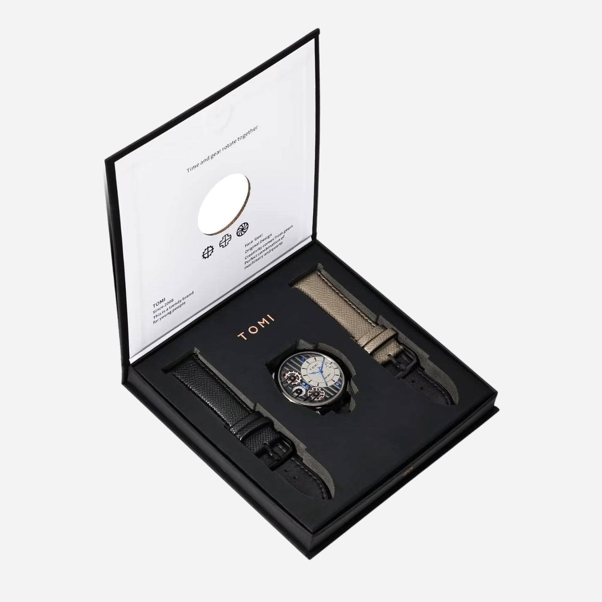 A luxury watch gift set by TOMI, featuring a central chronograph watch with a black and blue dial, a black leather strap, and a beige woven strap, presented in an open black gift box with a white interior insert.