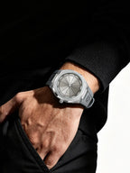 A close-up of a person's hand and wrist wearing a silver-toned watch with a matte gray dial and matching gray strap, styled with a black knit sweater against a dark background.