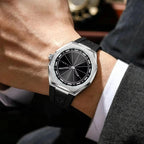 A close-up of a men's wristwatch with a silver-tone octagonal bezel, black dial, and black rubber strap, worn on a person's wrist with a white dress shirt cuff and gray suit jacket visible.