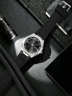 A wristwatch with a black strap and silver-toned case, photographed in a styled flat-lay with a flashlight, phone, and dark textured fabric around it.