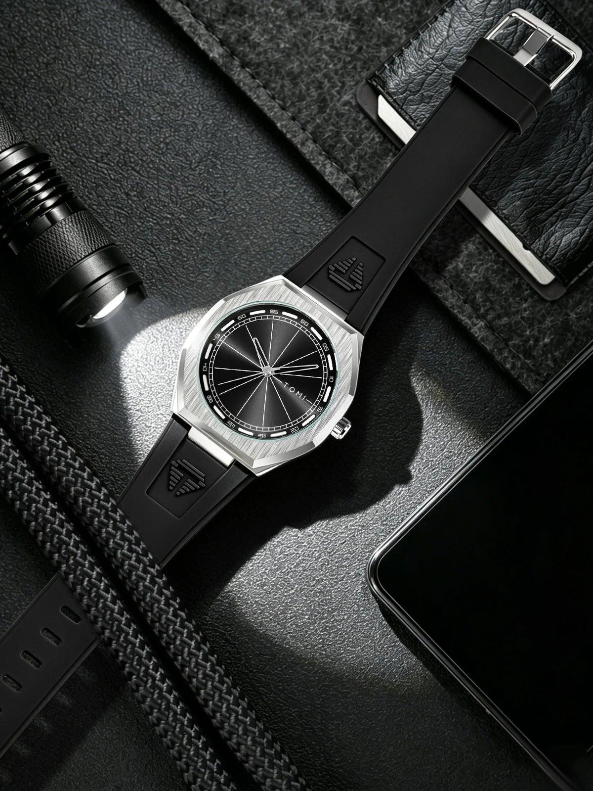 A wristwatch with a black strap and silver-toned case, photographed in a styled flat-lay with a flashlight, phone, and dark textured fabric around it.