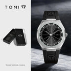 TOMI analog watch with a brushed silver-tone octagonal case, black dial, and black rubber strap, shown alongside its black presentation box on a split light gray background.