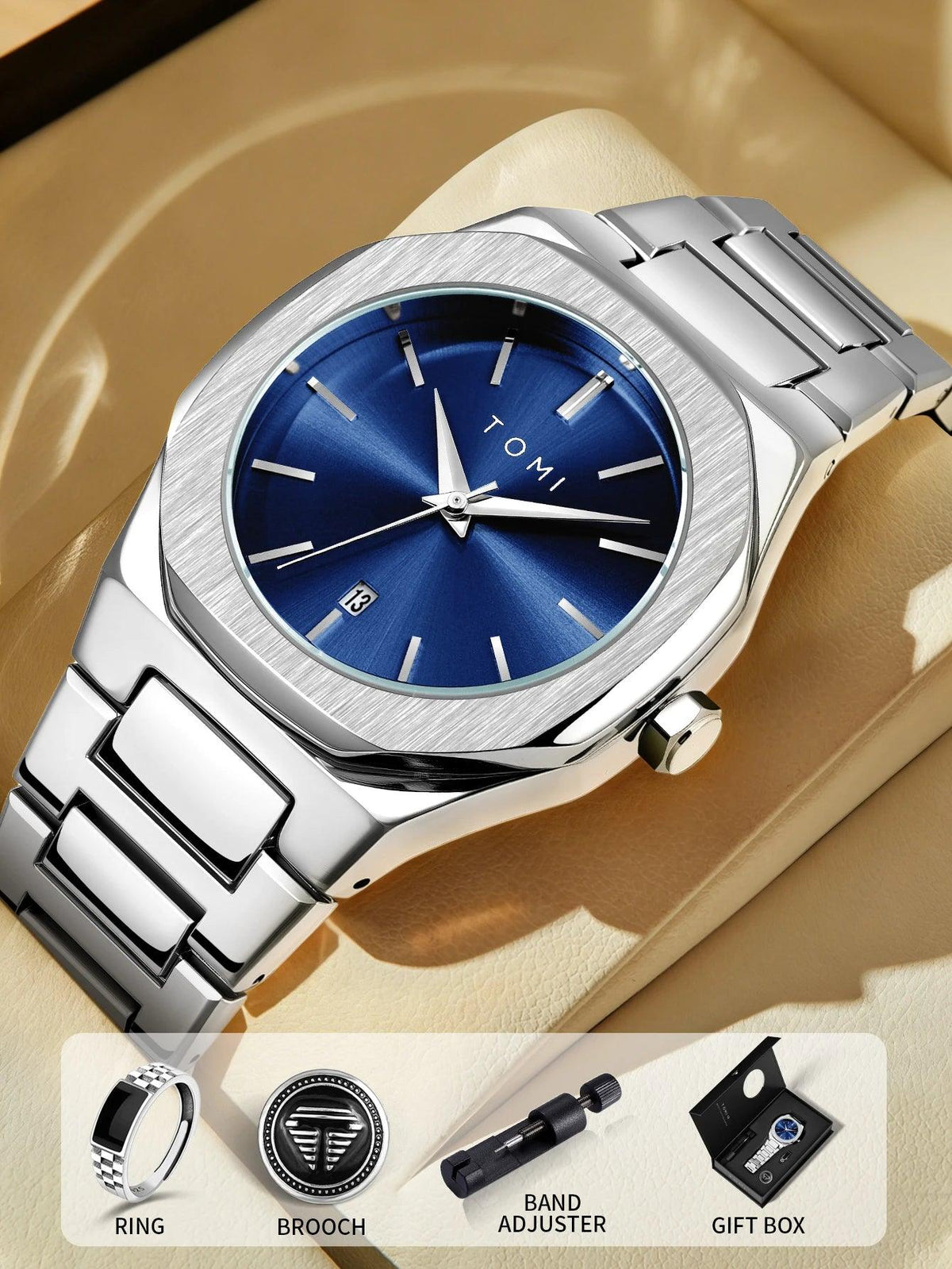 A silver stainless steel wristwatch with a blue sunburst dial and date window, displayed on a beige surface with a watch box. The image includes a bottom banner showing included accessories: a ring, brooch, band adjuster, and gift box.