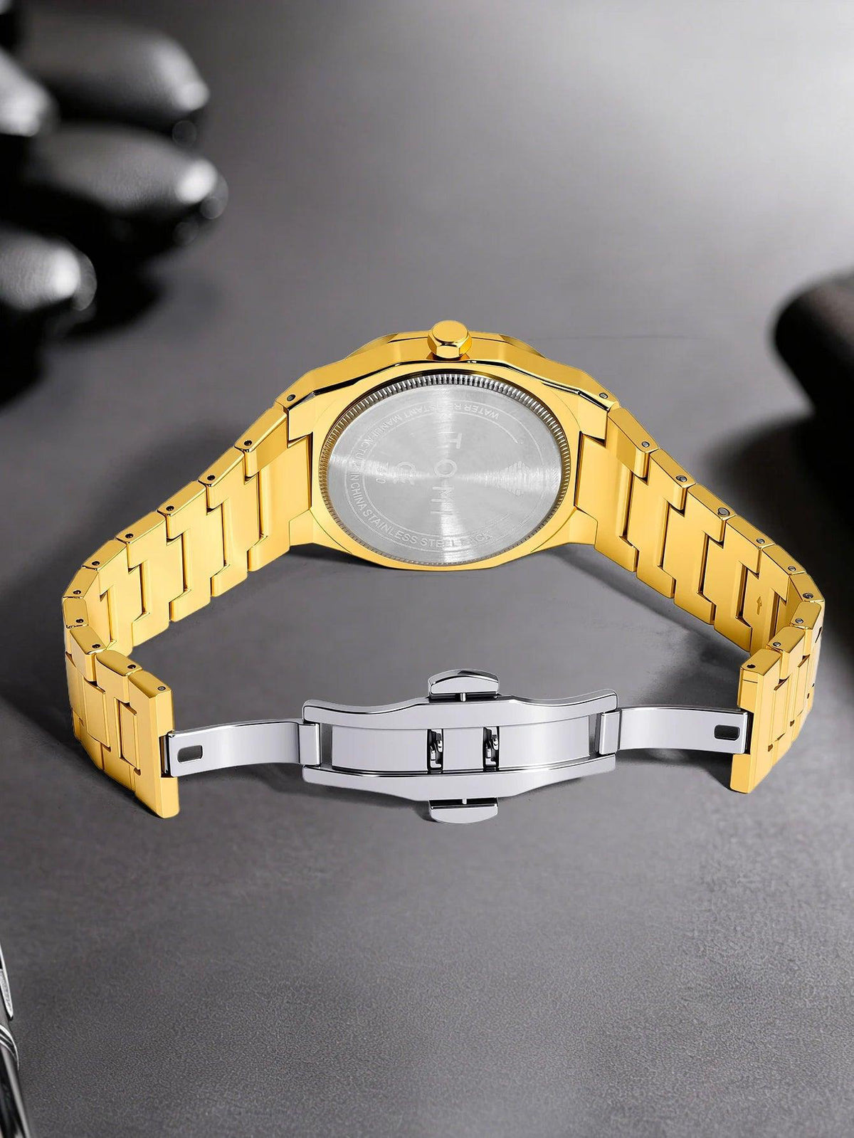 Gold-tone stainless steel watch with a matching metal link bracelet and a silver-toned folding clasp, photographed on a dark gray surface.