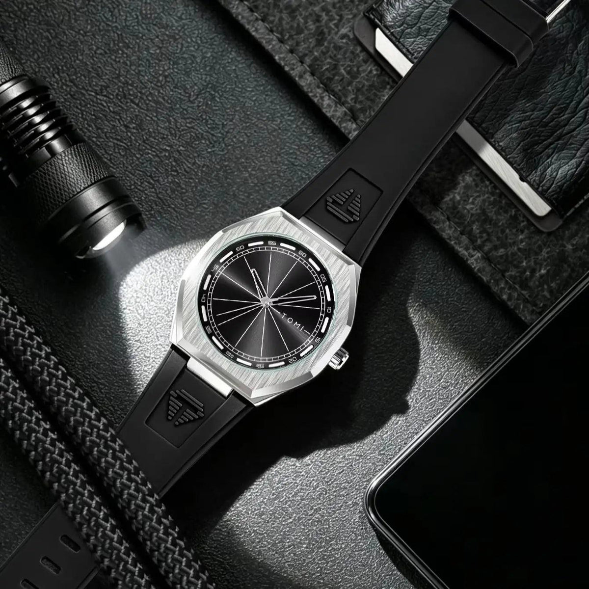 A black and silver wristwatch with a textured octagonal bezel and black strap, styled on a dark textured surface with a flashlight, braided strap, and smartphone nearby.