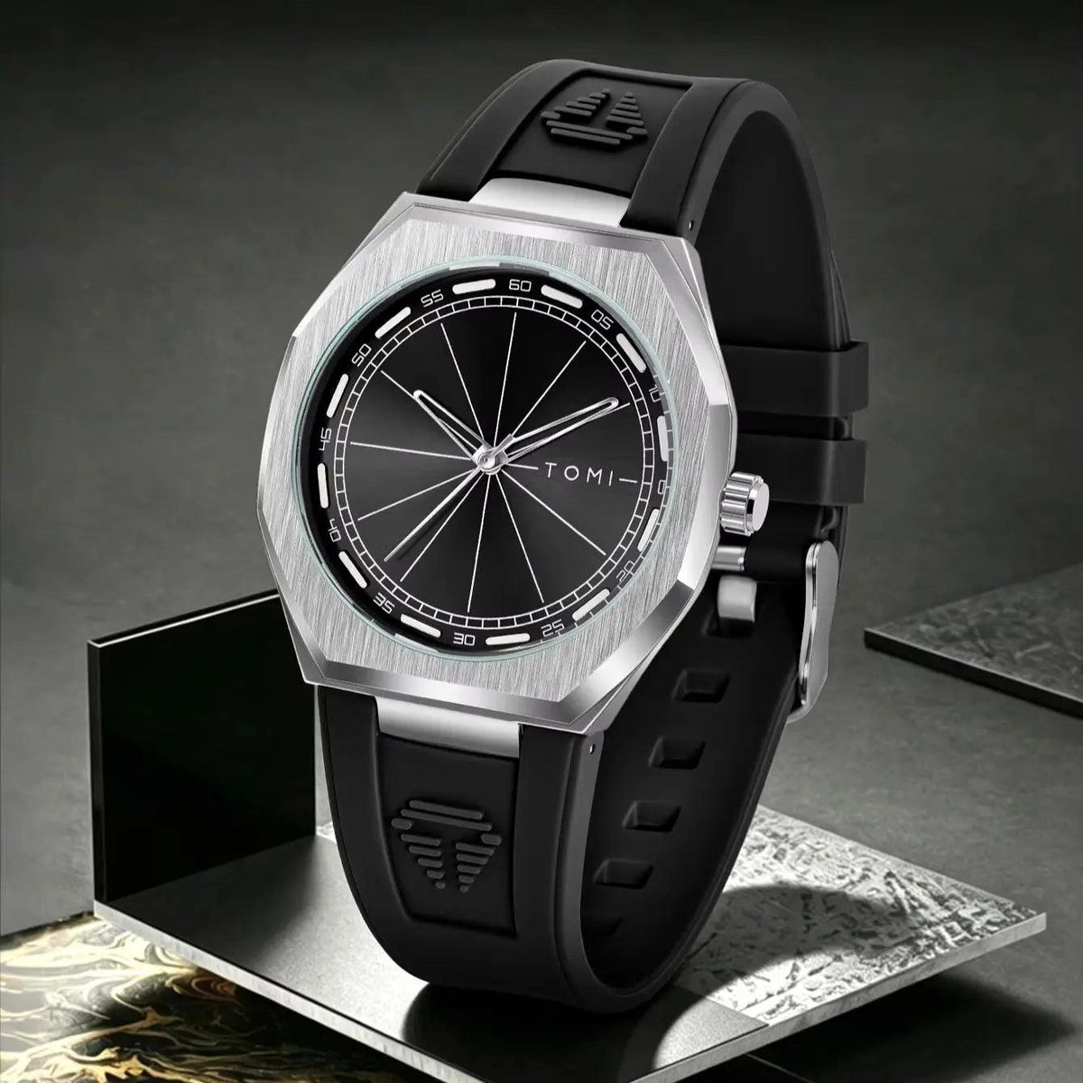 A TOMI men's watch with a brushed silver-tone octagonal bezel, black dial, and black rubber strap, displayed on a metallic stand against a dark gray background.