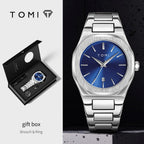 TOMI stainless steel watch with a blue dial and silver bracelet, shown alongside its black gift box and brooch and ring packaging on a split white and dark background.