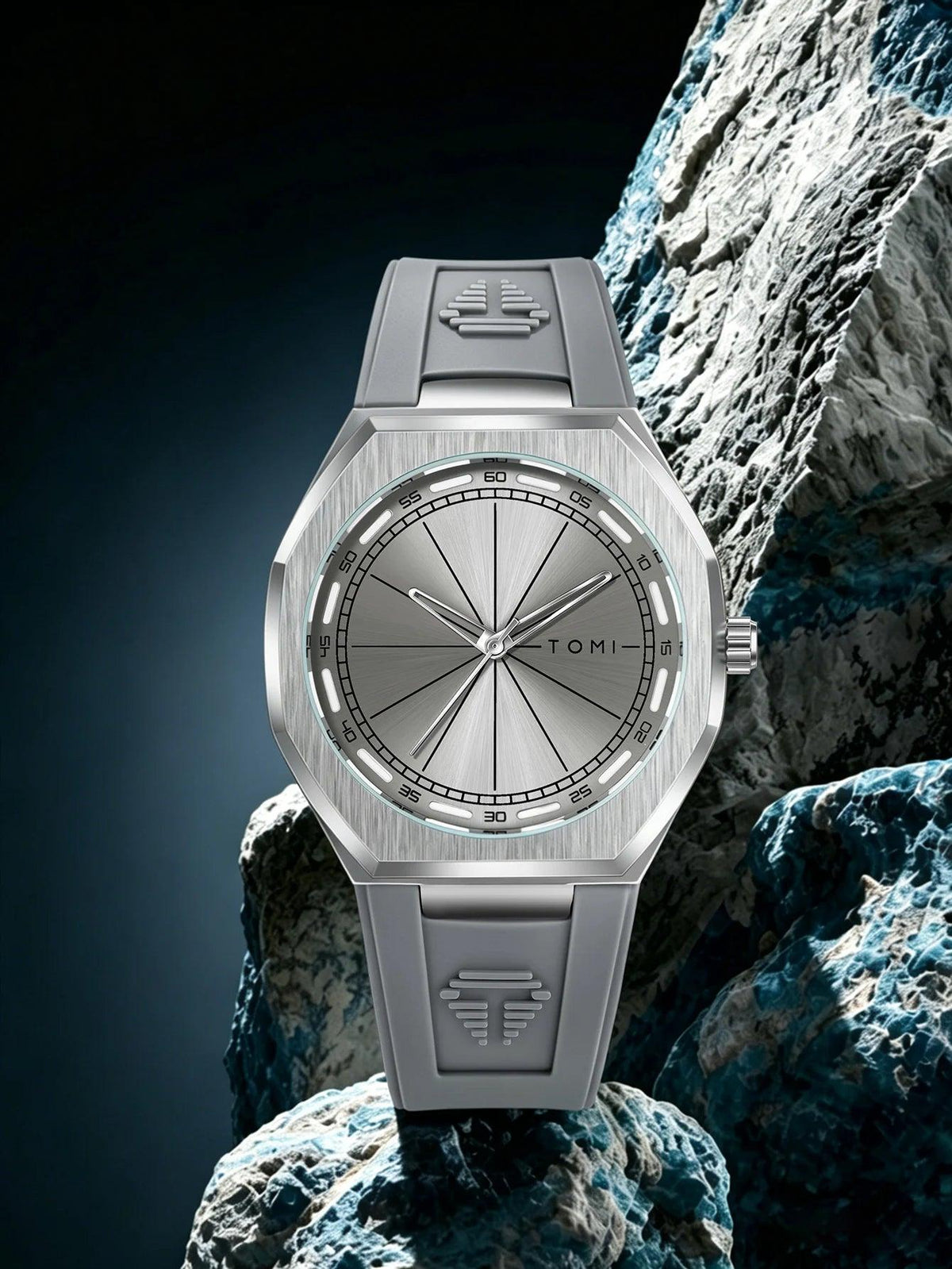 A silver-tone TOMI watch with a brushed metal case and matching dial, shown standing upright on textured blue-gray rocks against a dark studio background.