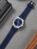A wristwatch with a dark blue dial and a dark blue silicone strap, laid diagonally on a textured gray fabric surface next to a black carbon-fiber-patterned case and a wooden block.