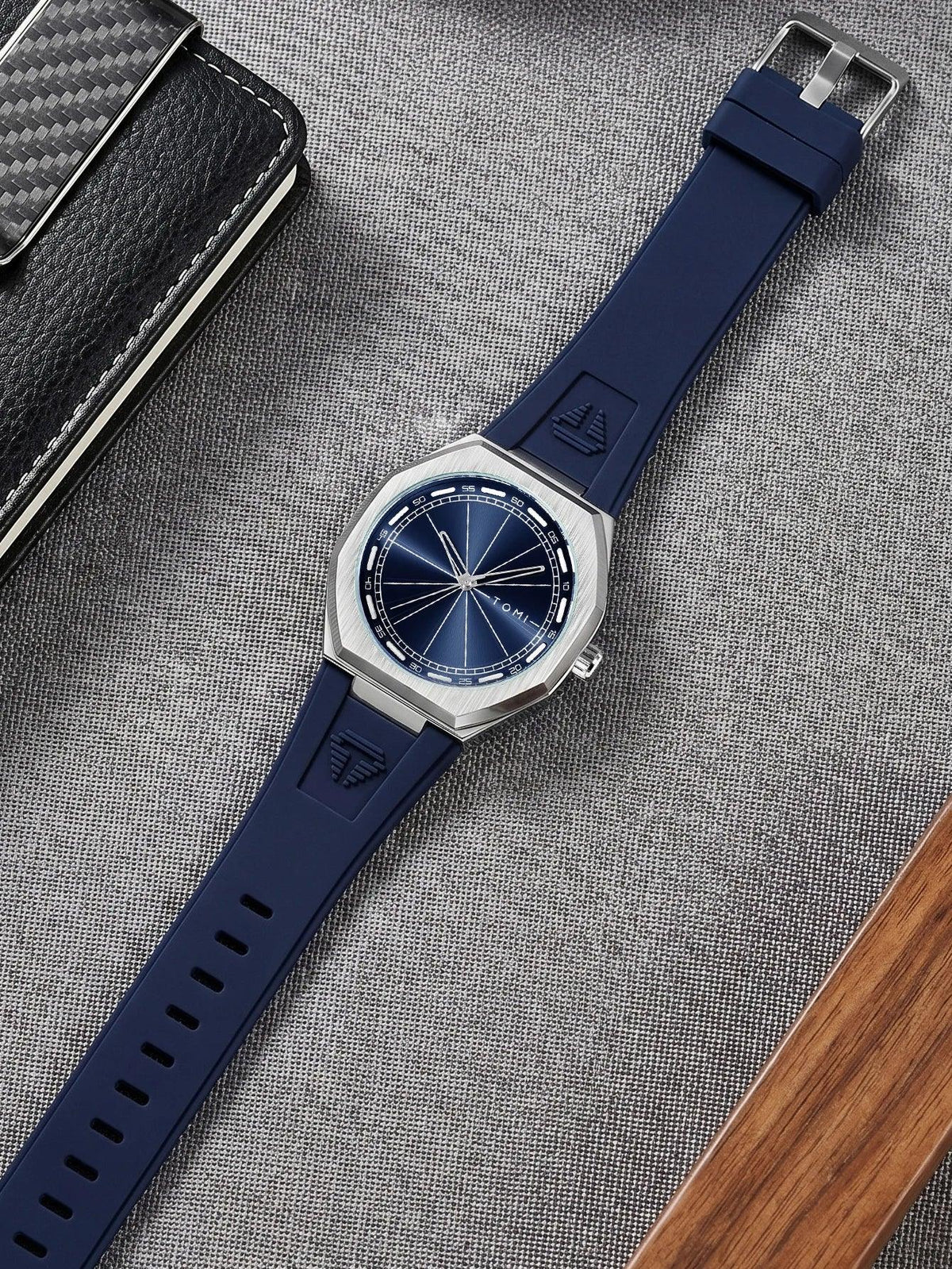 A wristwatch with a dark blue dial and a dark blue silicone strap, laid diagonally on a textured gray fabric surface next to a black carbon-fiber-patterned case and a wooden block.