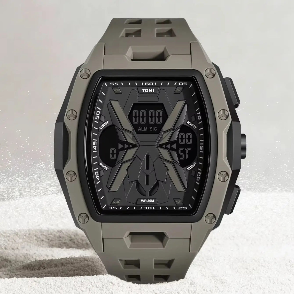 A rugged digital watch with a matte gray-green case and strap, featuring a black face with geometric graphics and multiple displays, set against a light gray background with subtle white specks.
