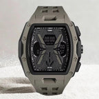 A rugged digital watch with a matte gray-green case and strap, featuring a black face with geometric graphics and multiple displays, set against a light gray background with subtle white specks.