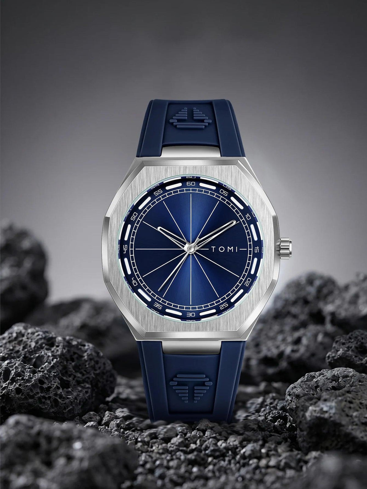 A TOMI watch with a brushed silver-tone case, deep blue dial, and blue silicone strap, displayed upright on dark volcanic rocks against a dark gray gradient background.
