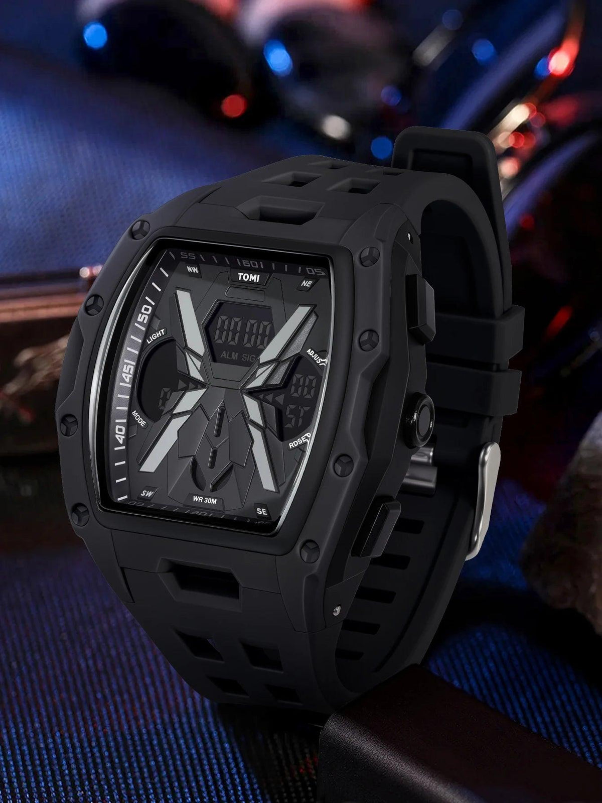 A rugged black digital sports watch with a thick protective case and matching black strap, shown close up against a dark blue and black textured background with blurred colorful bokeh.