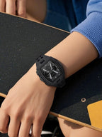 A black rugged analog watch with a textured dial and black strap worn on a person's wrist, resting on a black skateboard with a blue sleeve visible.