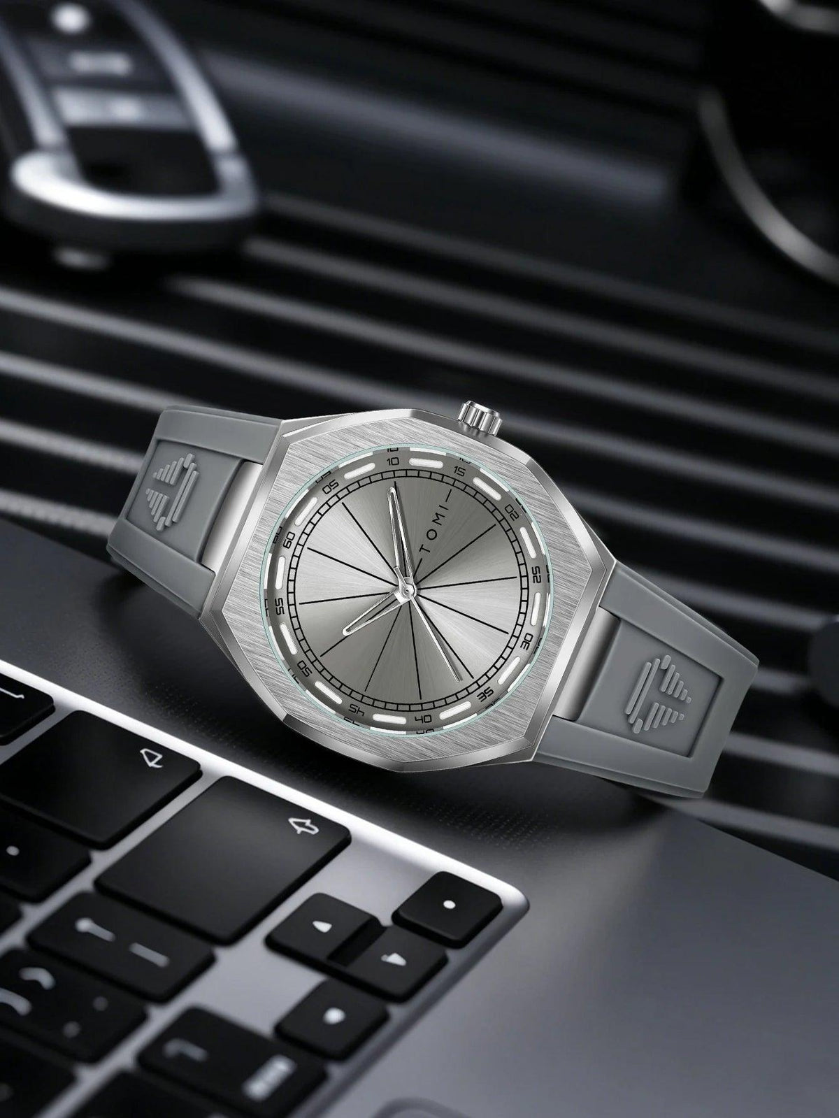 A silver-toned Tommi watch with a brushed metal case and strap, resting on a laptop keyboard with a blurred car key fob in the background.