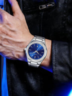 A silver-toned metal wristwatch with a deep blue dial and matching blue hands, shown worn on a person's wrist against a black leather jacket background.