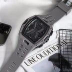 A rugged gray digital watch with a textured strap and black face, styled on a white fabric surface next to a smartphone, a magazine, and a black wire rack.