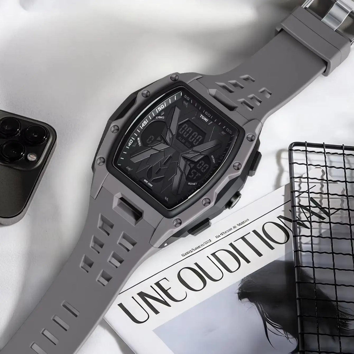 A rugged gray digital watch with a textured strap and black face, styled on a white fabric surface next to a smartphone, a magazine, and a black wire rack.