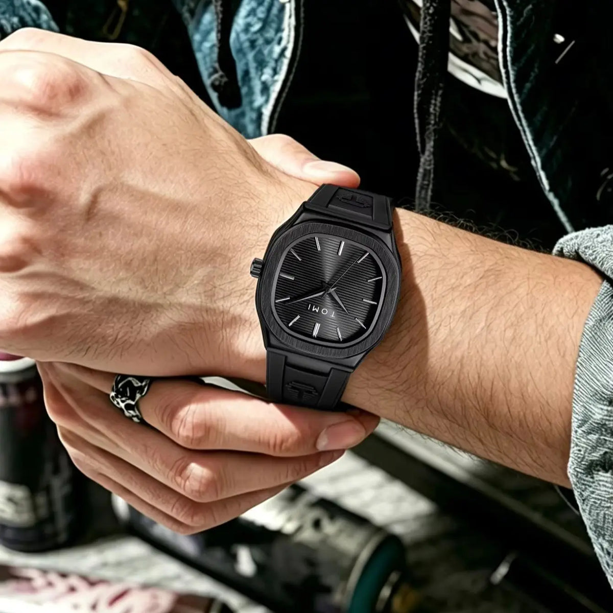 A close-up lifestyle shot of a man wearing a black rectangular analog watch with a textured sunburst dial and matte black strap. The watch is shown on his wrist, with his other hand resting on his knee.