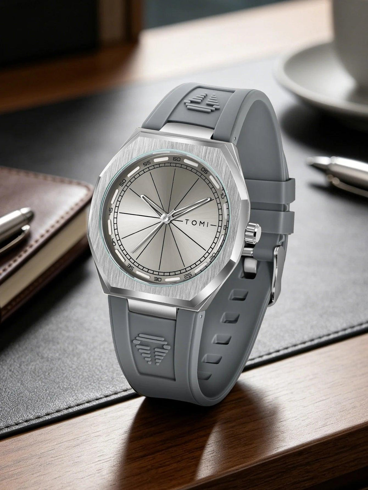 A silver and gray Tomi watch with a brushed metal case and a gray silicone strap, resting on a dark desk mat over a wooden surface.