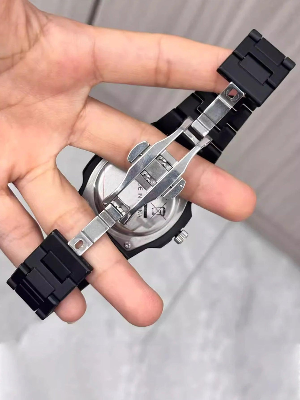 A close-up of a hand holding the back of a black and silver watch, showing the metal clasp and case details.