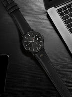 Black leather watch on a dark surface with a laptop and phone in the background