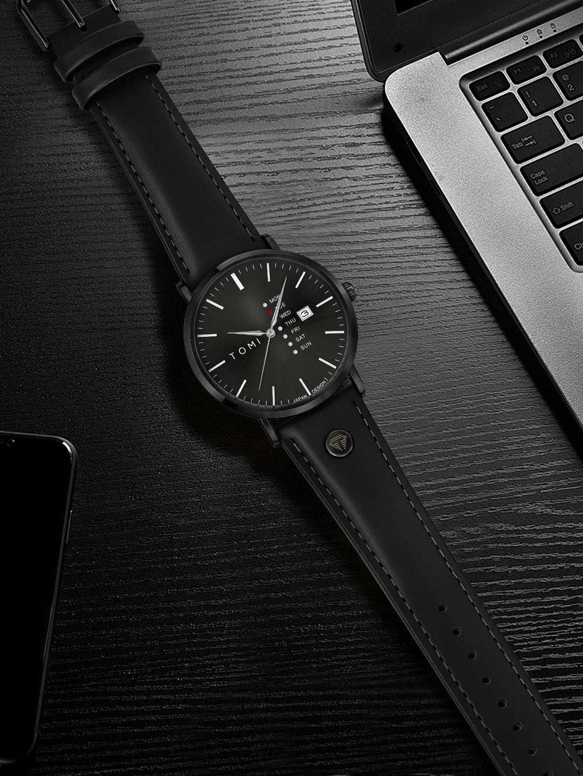 Black leather watch on a dark surface with a laptop and phone in the background