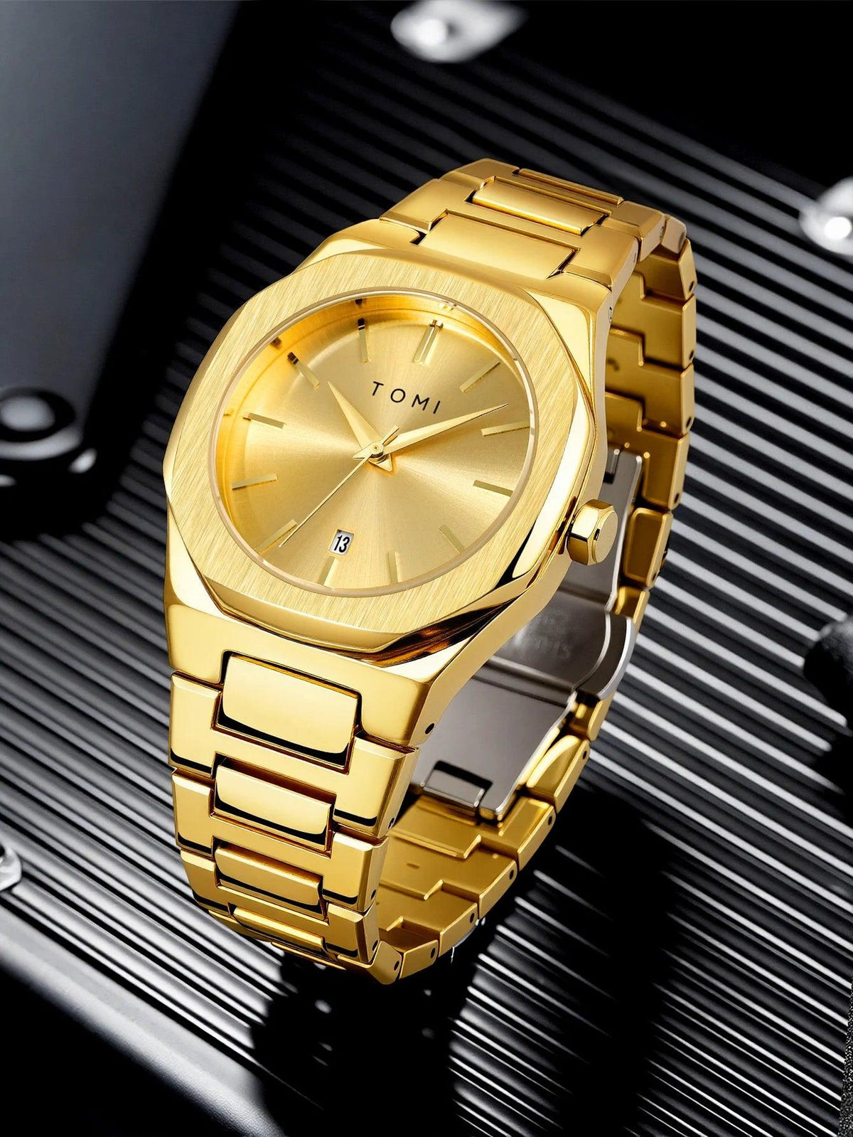 A gold-tone stainless steel wrist watch with a matching gold dial and link bracelet, resting on a black ribbed surface. The brand name 'TOMI' is visible on the dial.