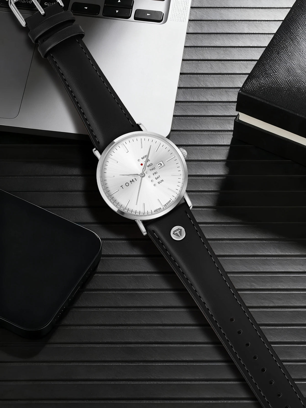 Black leather watch on a wooden surface with a smartphone and notebook.