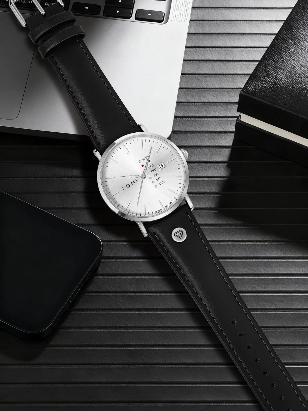 Black leather watch on a wooden surface with a smartphone and notebook.