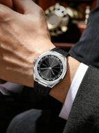 A close-up of a wrist wearing a silver-toned Tudor watch with a black dial and black rubber strap, shown on a person's wrist with a suit jacket and white dress shirt visible.