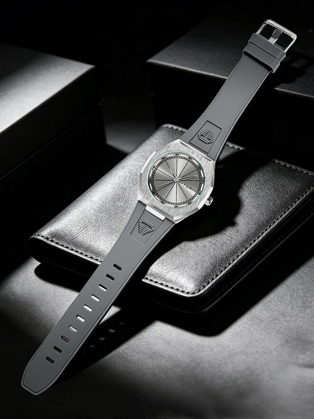A wristwatch with a dark gray strap and a silver-toned octagonal case, displayed diagonally on a textured black leather surface with dark background boxes.
