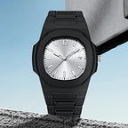 A black metal watch with a silver sunburst dial, shown resting on a textured stone surface against a light blue sky background.