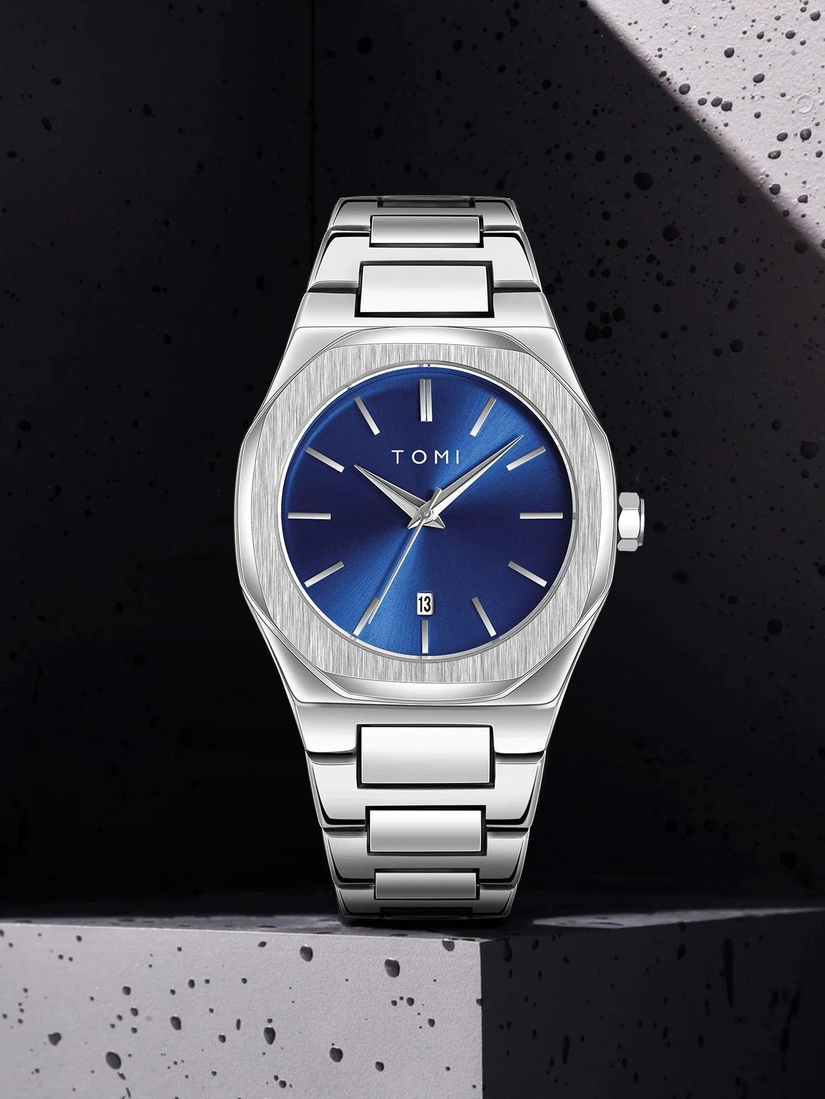 A TOMI men's watch with a brushed silver stainless steel case and bracelet, featuring a vibrant blue dial with silver-tone hands and indices, set against a dark textured background.